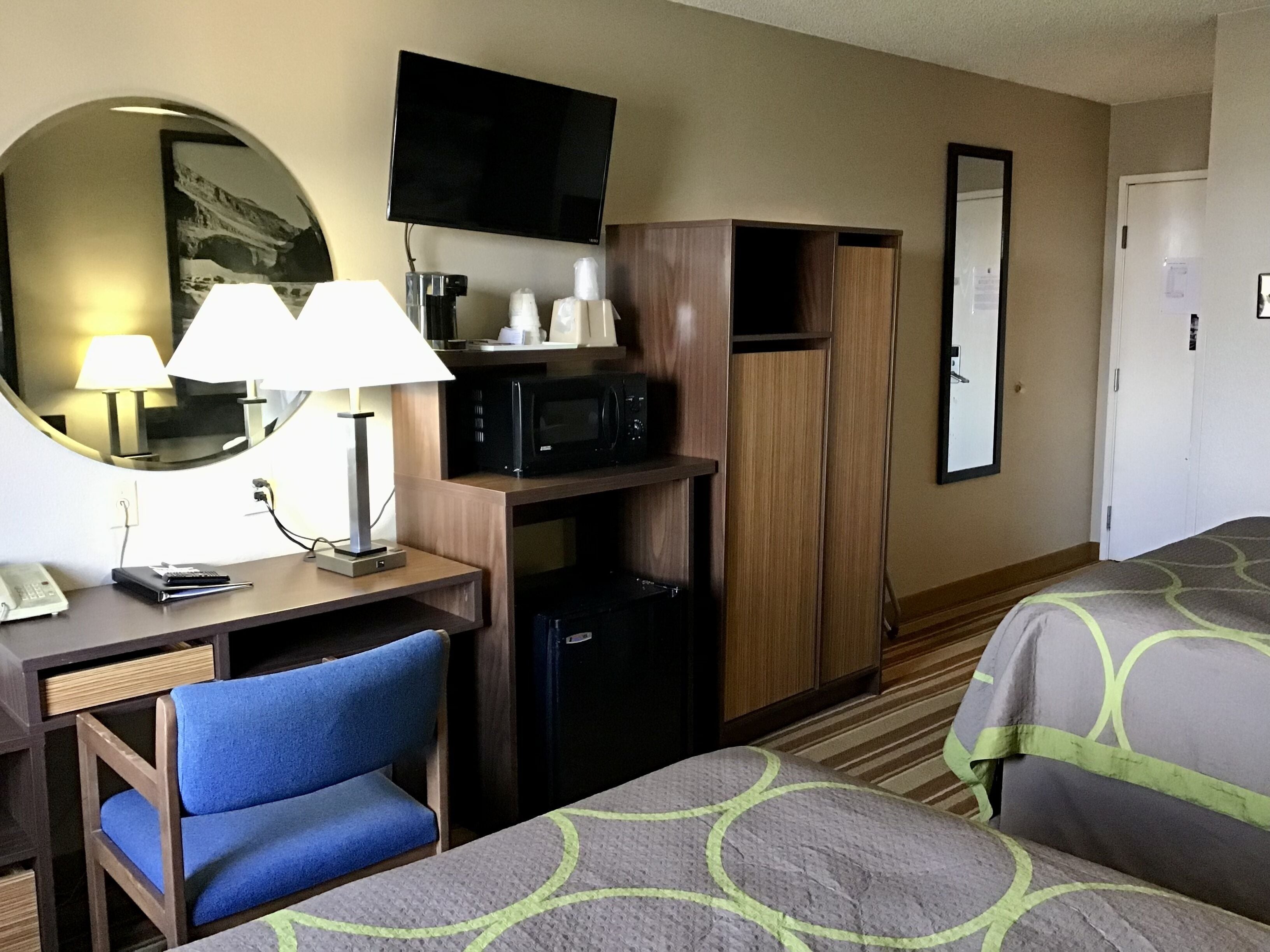 Ein Hotelzimmer mit Schreibtisch, zwei Stühlen, einer Mikrowelle, einem Kühlschrank, einem Fernseher und einem Bett mit gemusterten Bettbezügen.