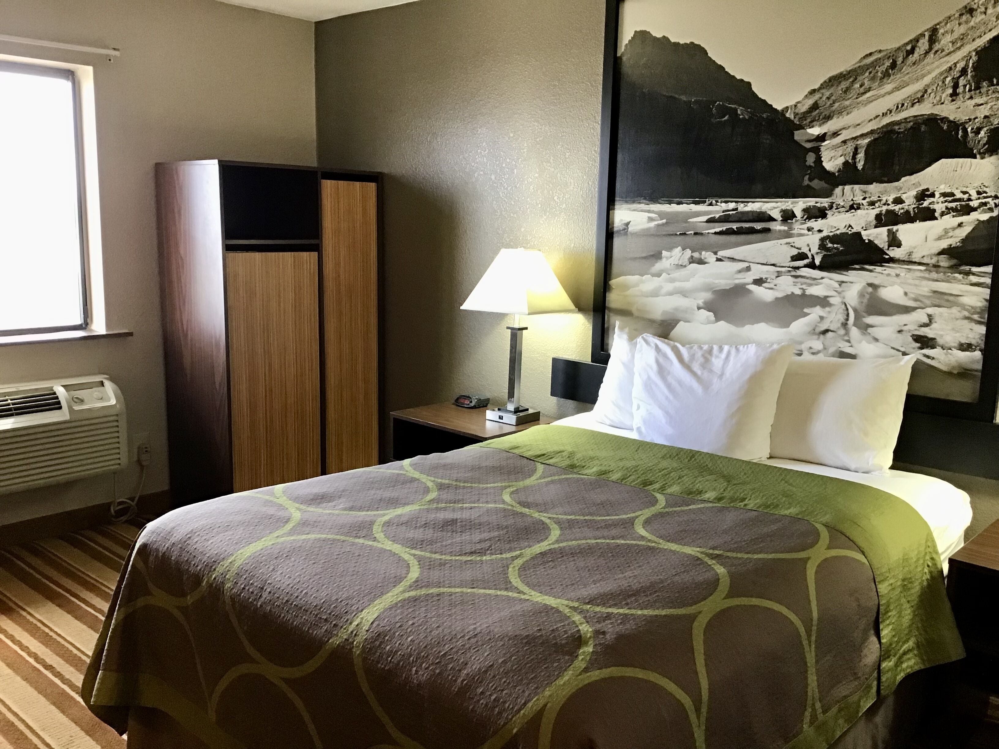 Habitación de hotel con cama, mesita de noche, lámpara, armario y un cuadro grande en la pared.