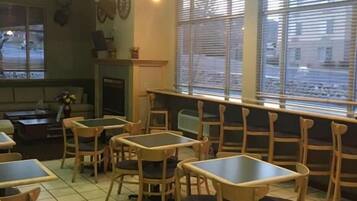 Interior de restaurante com mesas e cadeiras de madeira, lareira e amplas janelas com persianas.