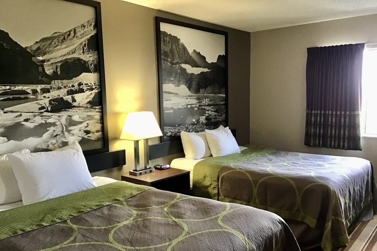 A hotel room with two beds, a nightstand, a lamp, and two framed pictures of mountains.