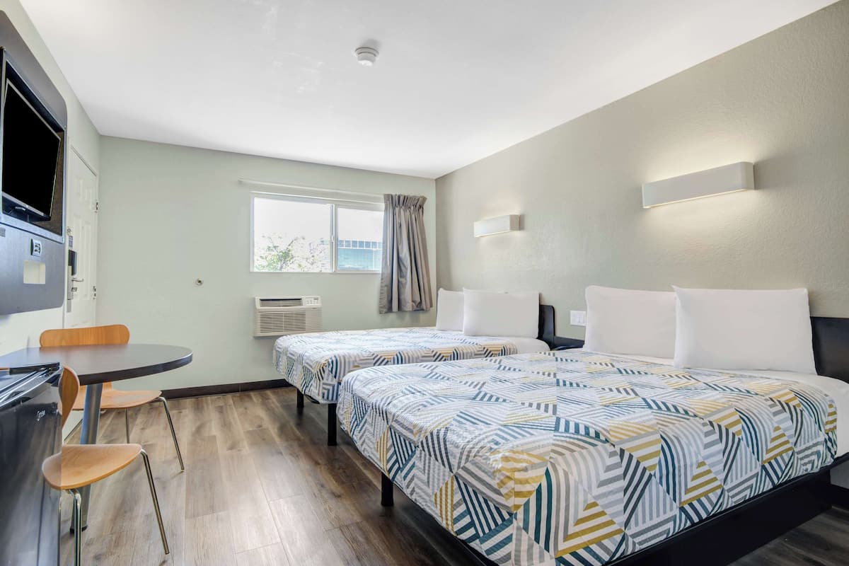 A hotel room with two beds, a desk, and a chair. There is a window with curtains, a flat-screen TV, and a wall-mounted light fixture.