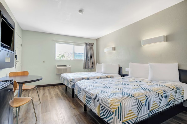 A hotel room with two beds, a desk, and a chair. There is a window with curtains, a flat-screen TV, and a wall-mounted light fixture.
