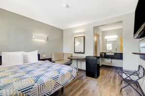 Una habitación de hotel con cama, escritorio, silla, refrigerador pequeño, lavamanos y espejo.