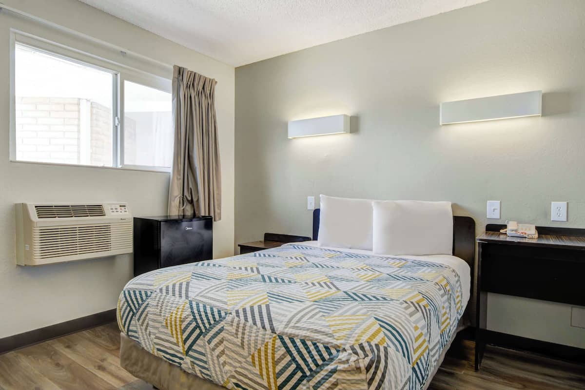 A hotel room with a bed, a nightstand, a lamp, an air conditioning unit, a window with curtains, and a wall-mounted light fixture.