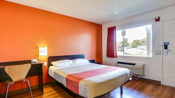 A hotel room with a bed, a desk with a chair, a window with curtains, and a door.