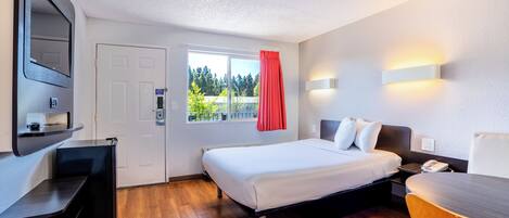A hotel room with a bed, bedside table, a desk, a chair, a window with a view, and a door.