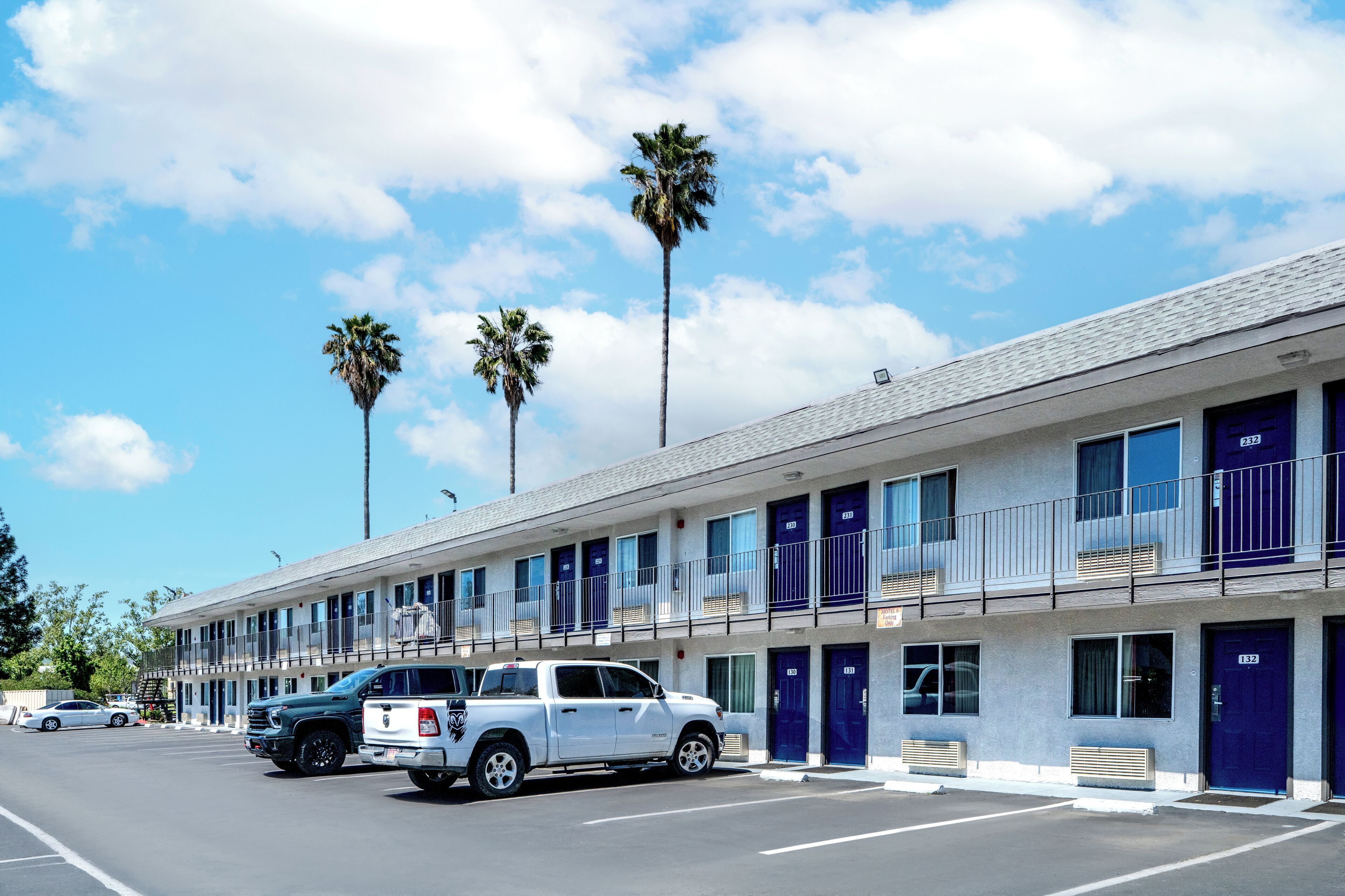 Un motel con una fila de habitaciones, cada una con puerta azul, balcón y estacionamiento con varios vehículos.