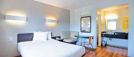 Una camera d'albergo con un letto, comodino, sedia e bagno con lavandino e specchio.