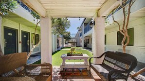 A covered outdoor area with wicker furniture, a small table with flowers, and a view of a building with numbered doors.