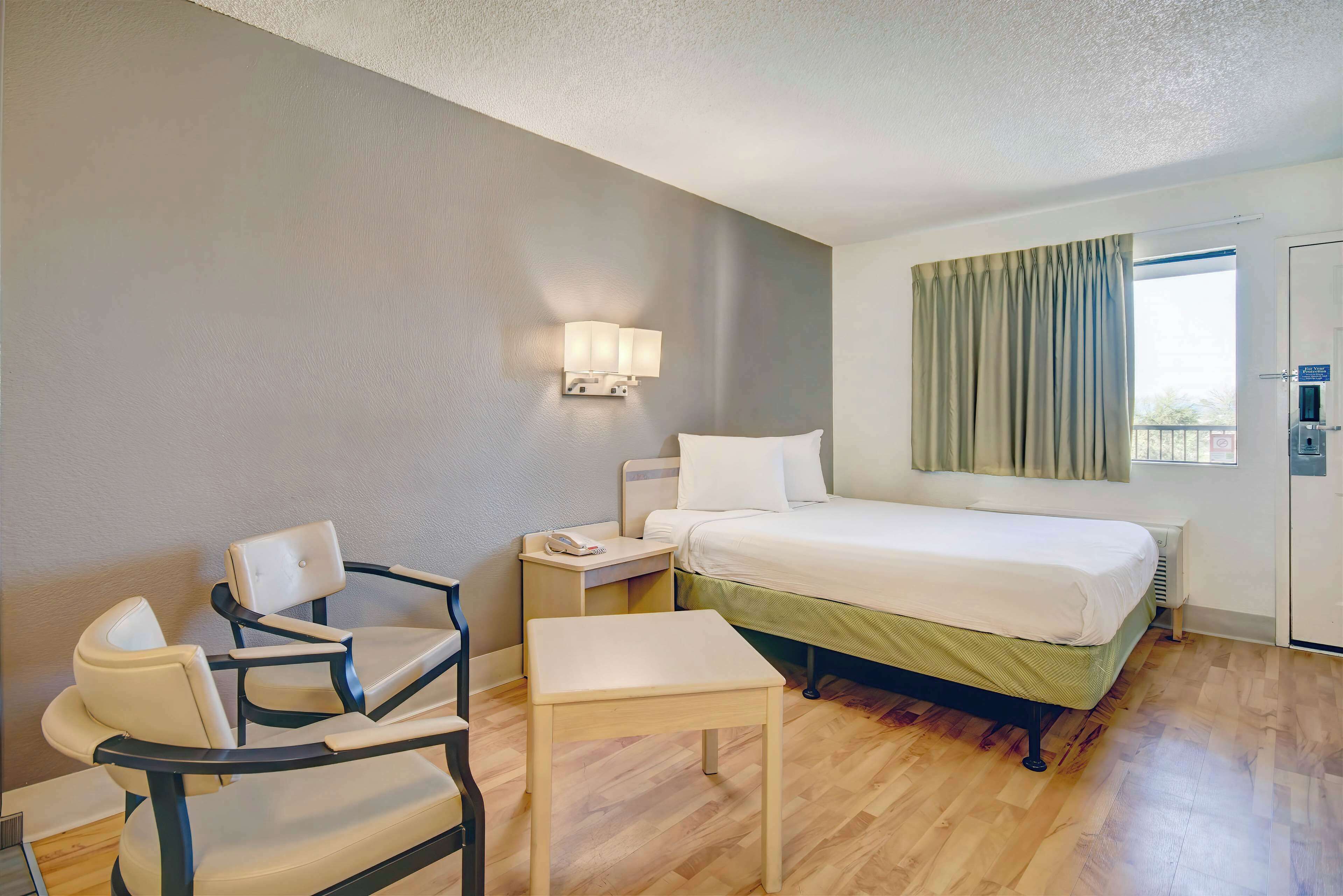 A hotel room with a bed, a desk, a chair, a nightstand, a lamp, and a window with curtains.