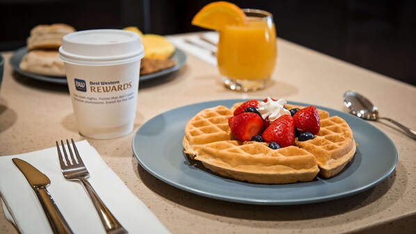 Un plato de desayuno con gofres, fresas, arándanos y crema batida, una taza de café y un vaso de jugo de naranja sobre una mesa.