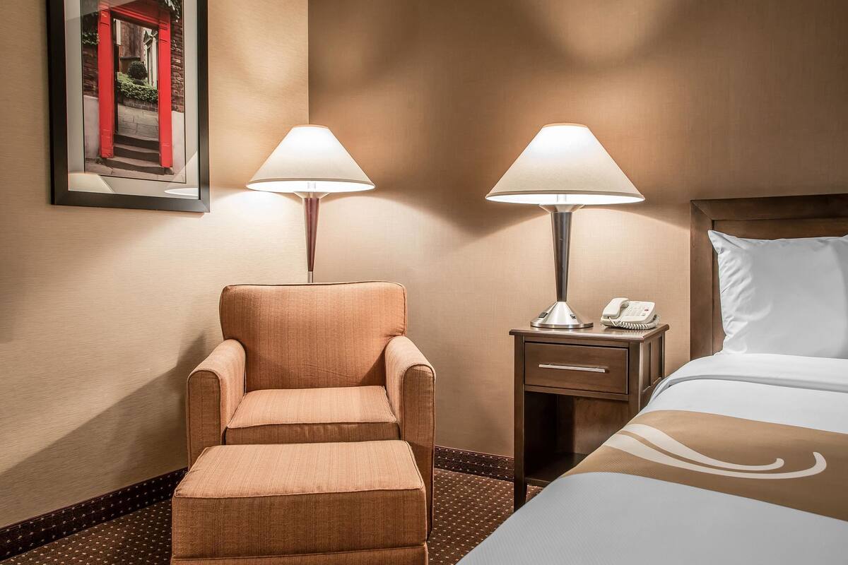 A hotel room with a bed, a chair, a nightstand with a lamp, and a framed picture on the wall.