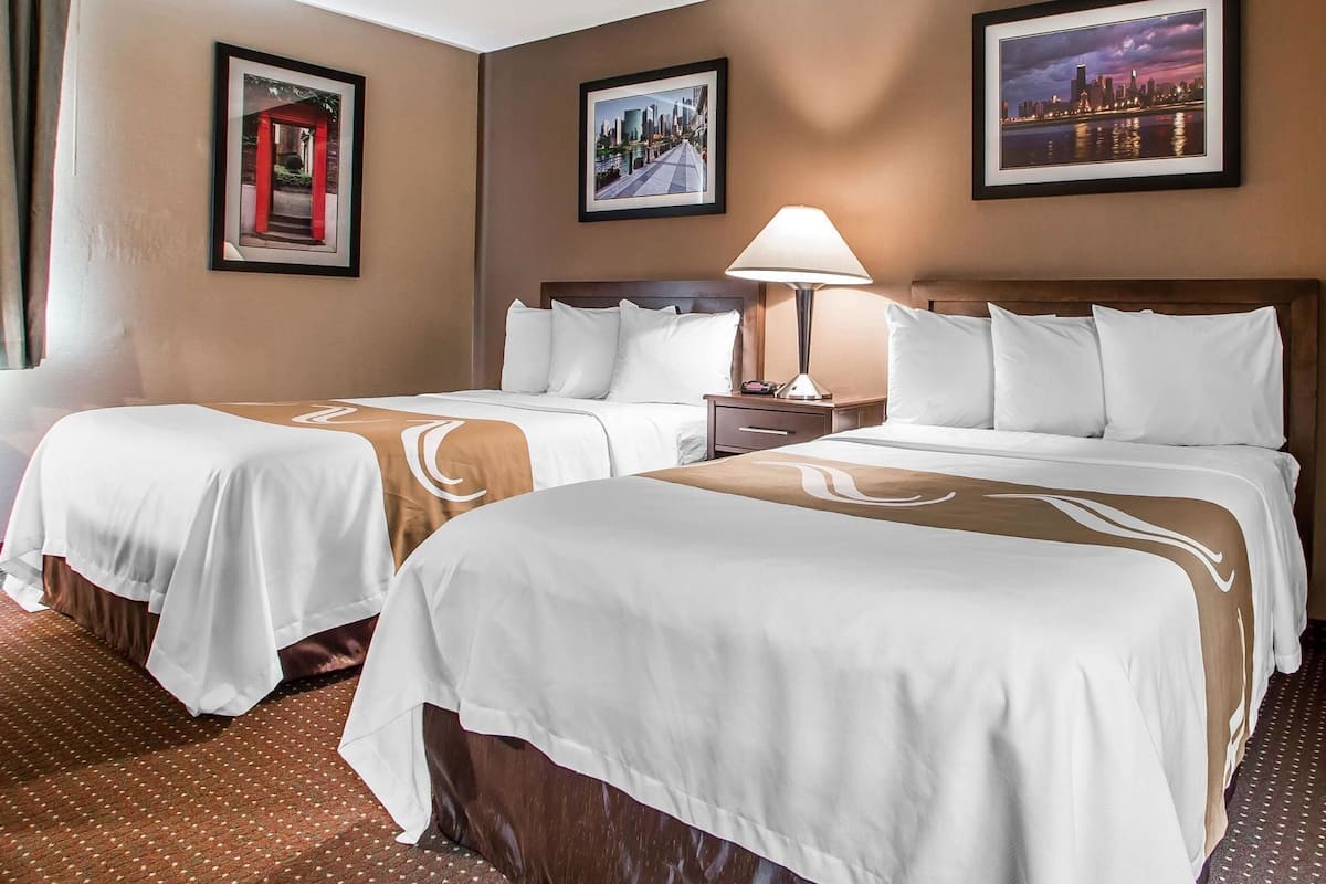 A hotel room with two beds, a nightstand, a lamp, and framed pictures on the walls.