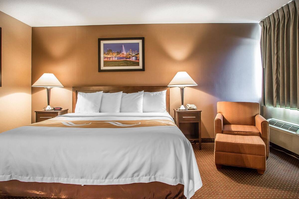 A hotel room with a bed, two bedside lamps, a chair, a picture on the wall, and a window with curtains.