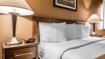 Habitación de hotel con cama, dos lámparas de noche, mesita de noche con reloj y un cuadro en la pared.