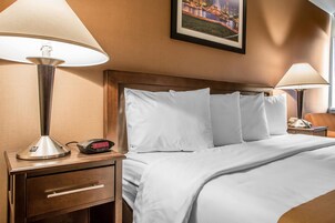 Habitación de hotel con cama, dos lámparas de noche, mesita de noche con reloj y un cuadro en la pared.