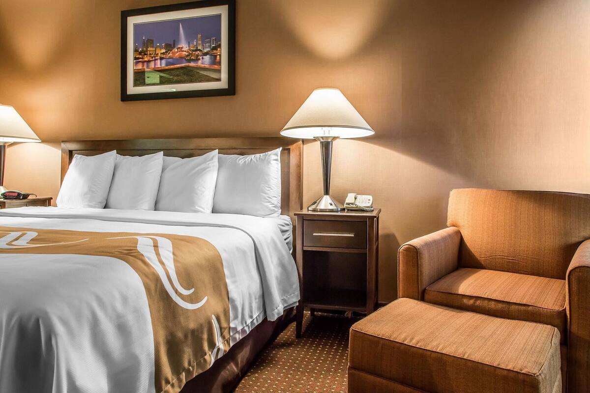 A hotel room with a bed, two bedside lamps, a nightstand, a chair, and a framed picture on the wall.