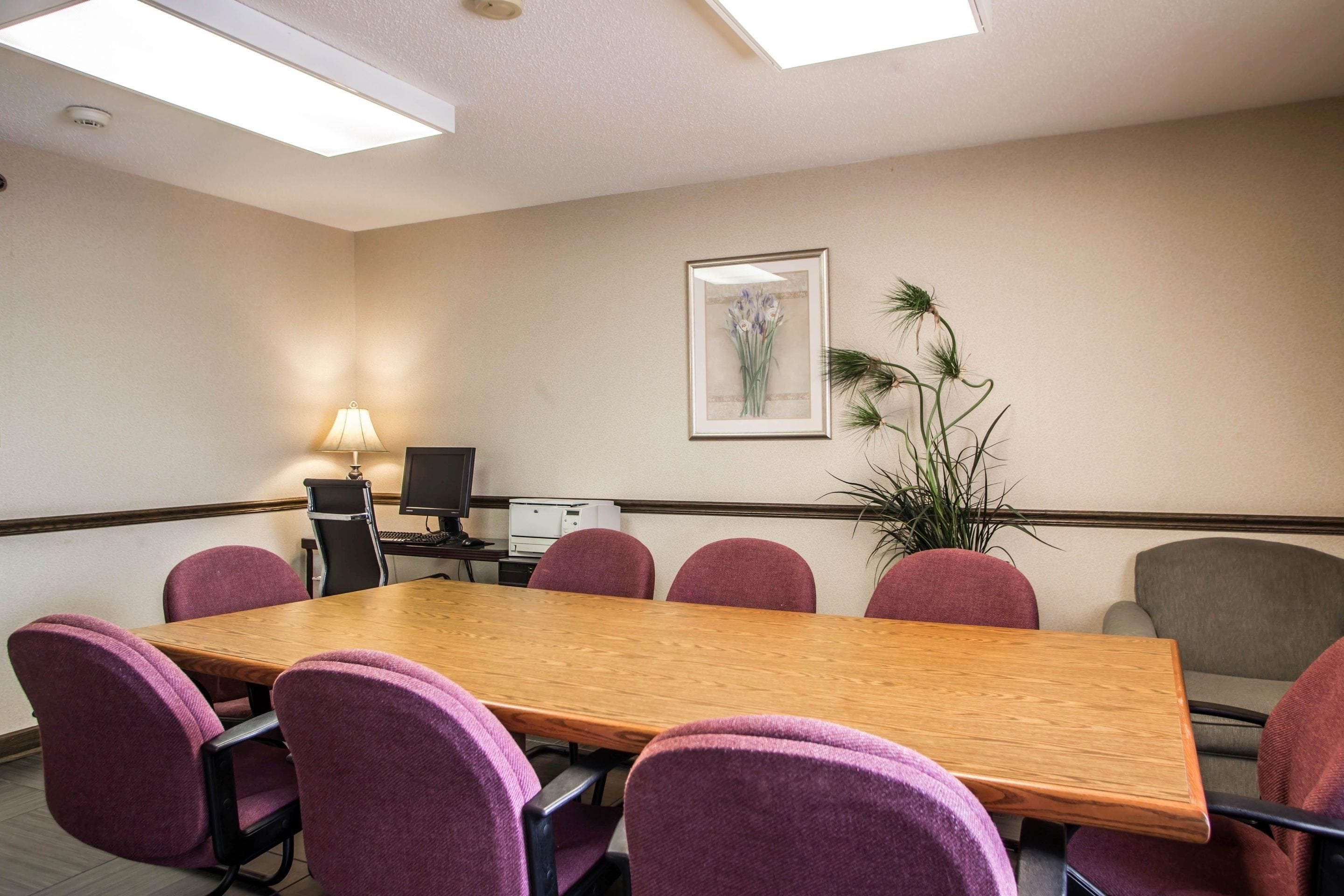 Un salón de conferencias con una larga mesa de madera, sillas moradas, un escritorio con una lámpara, una computadora y un cuadro en la pared.