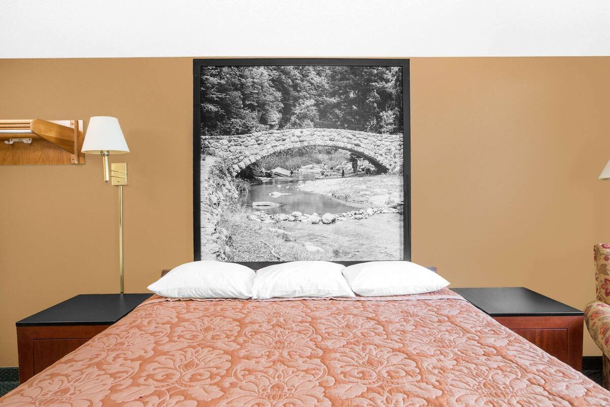A hotel room with a bed, a nightstand, a lamp, and a framed black and white photograph of a stone bridge over a river.