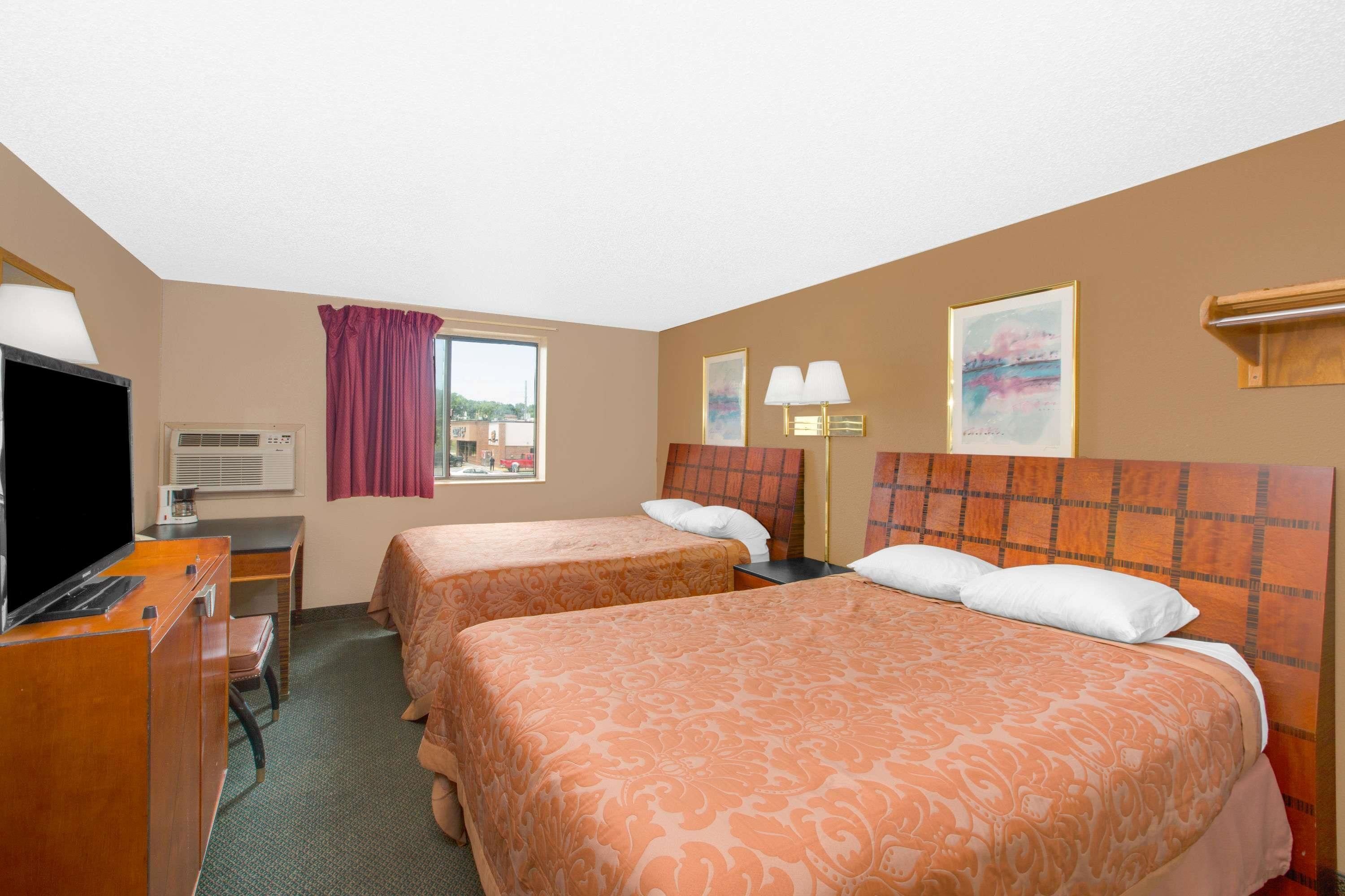 A hotel room with two beds, a desk with a television, an air conditioning unit, and artwork on the walls.