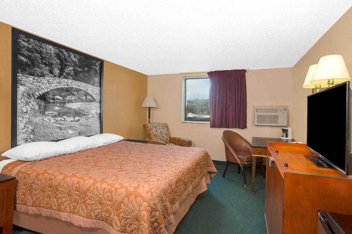 A hotel room with a bed, a desk with a television, a chair, a window with curtains, and a wall-mounted air conditioning unit.
