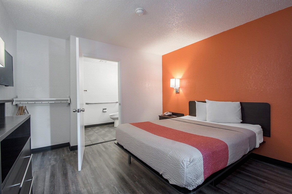 A hotel room with a bed, a nightstand, a wall-mounted lamp, a door leading to a bathroom, and a small desk with a chair.