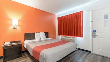 A hotel room with an orange wall, a bed with white and red bedding, a nightstand, a window with red curtains, and a door with a safety sign.