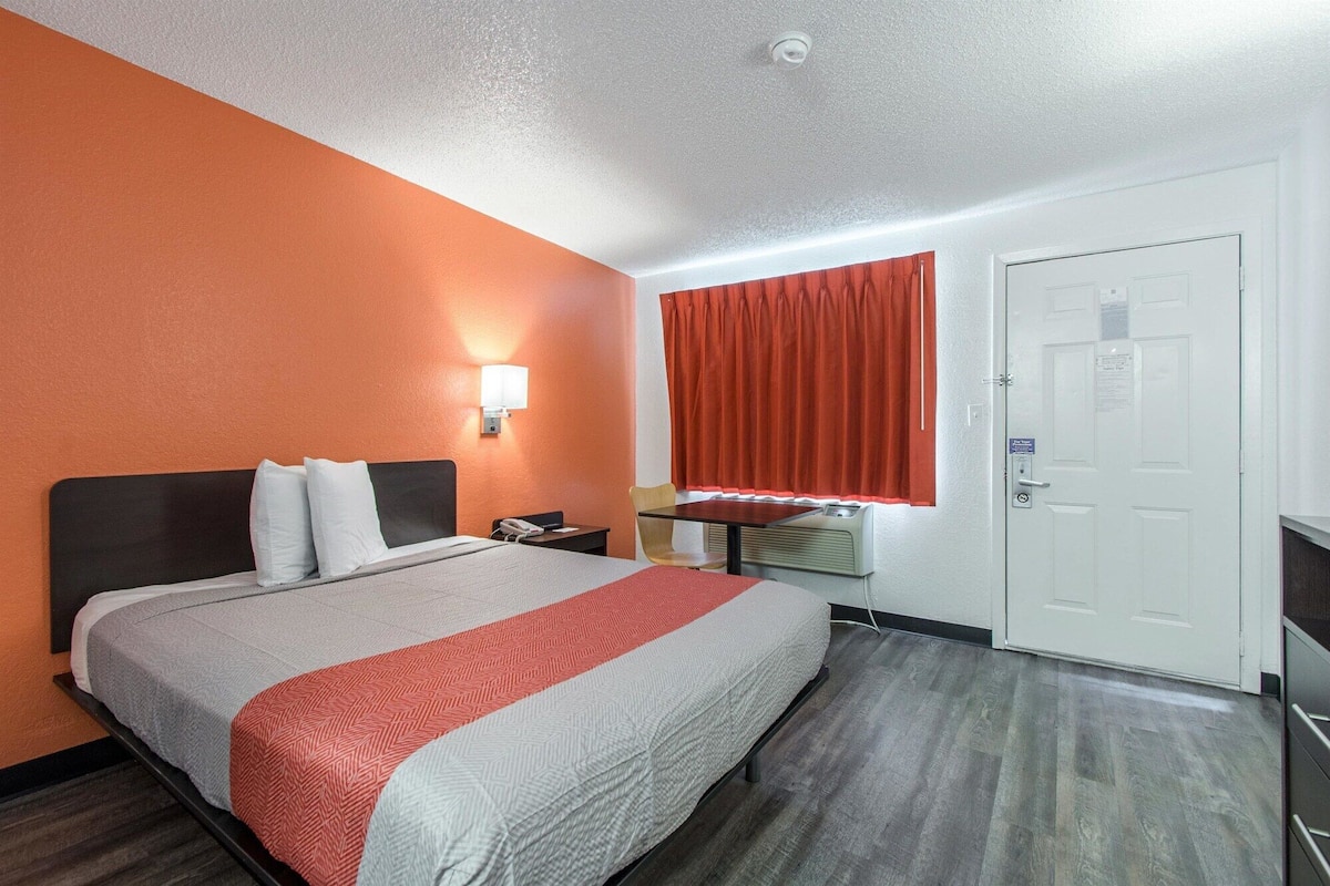 A hotel room with a bed, a desk, a chair, a window with curtains, and a door.