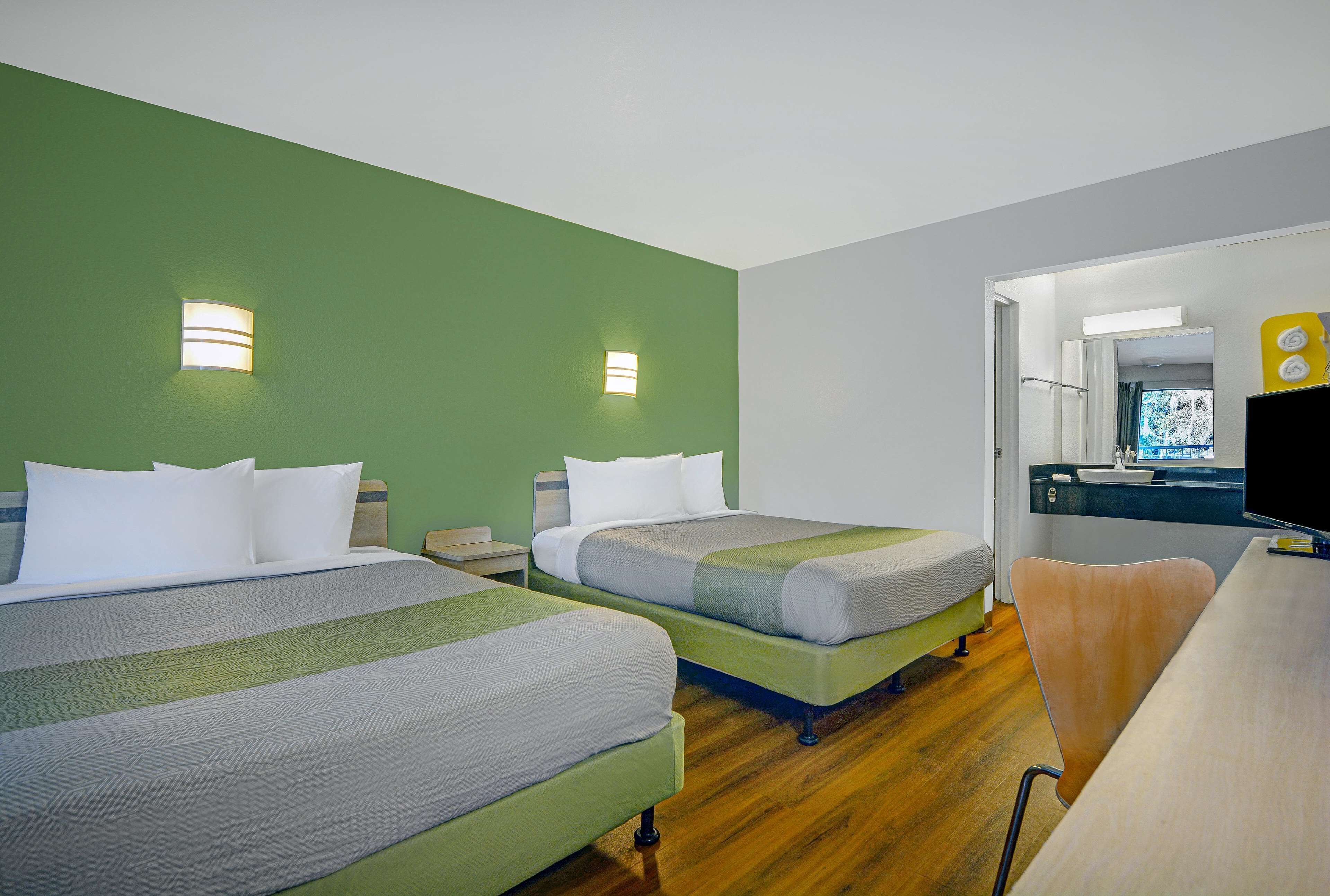A hotel room with two beds, a desk, a chair, a television, and a bathroom visible through an open door.