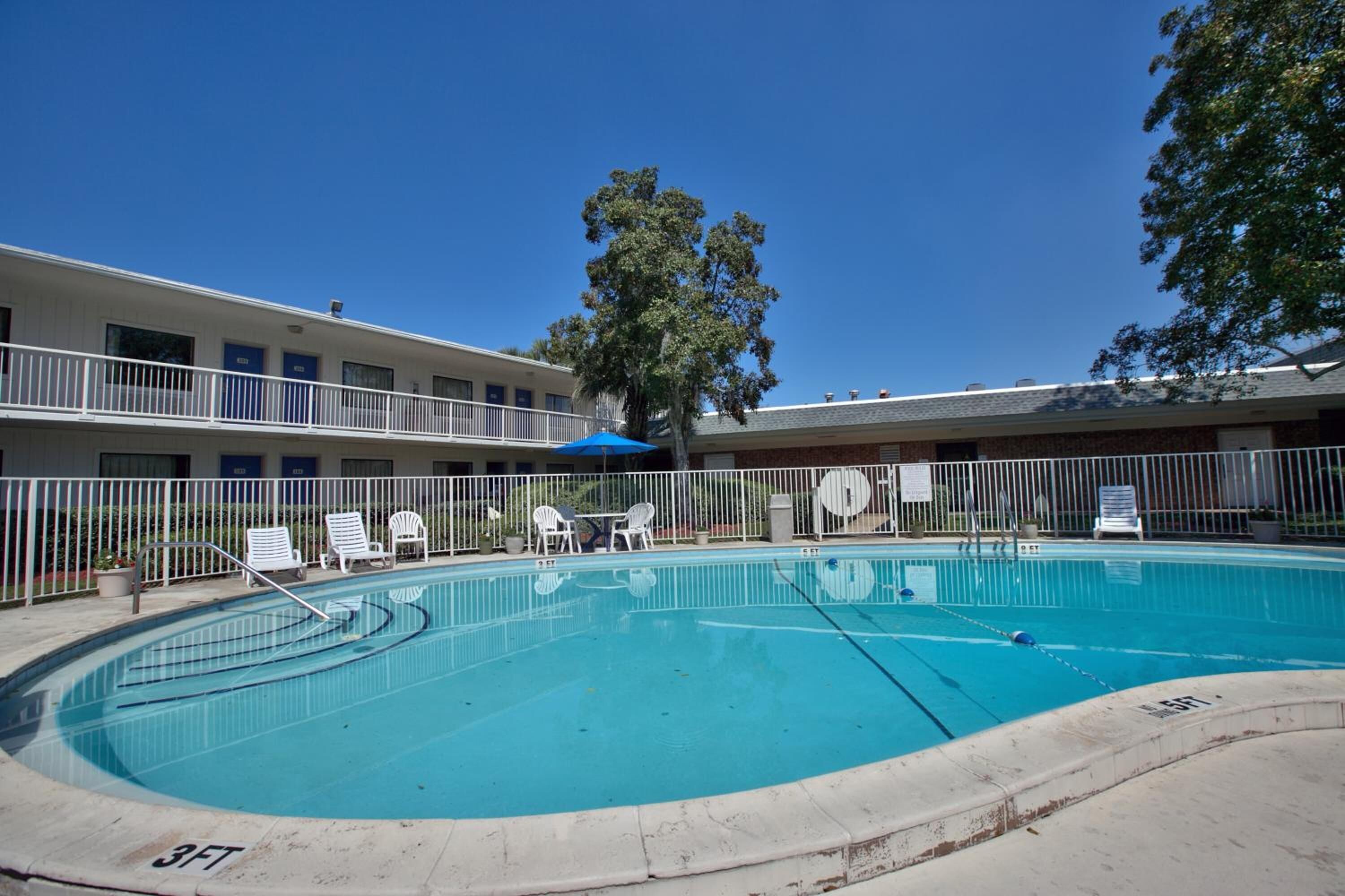 A swimming pool with lounge chairs and a blue umbrella, surrounded by a white fence and a building with multiple windows.