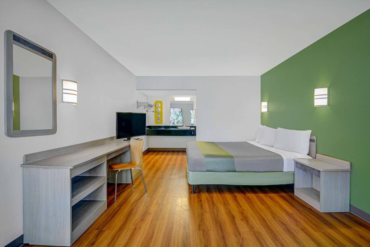 A modern hotel room with a wooden floor, a bed with white bedding, a desk with a chair, a TV, and a green accent wall.