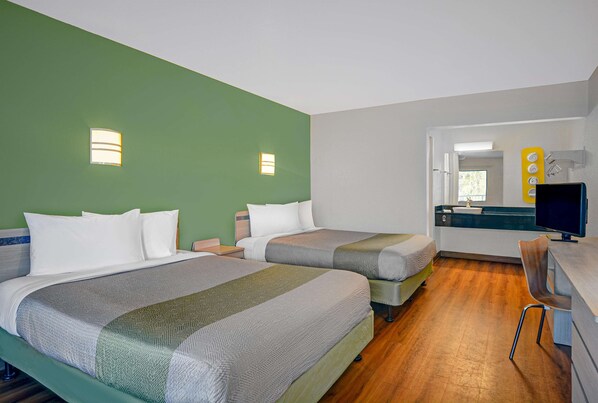 Habitación de hotel con dos camas, un escritorio con computadora y un baño visible a través de una puerta abierta.