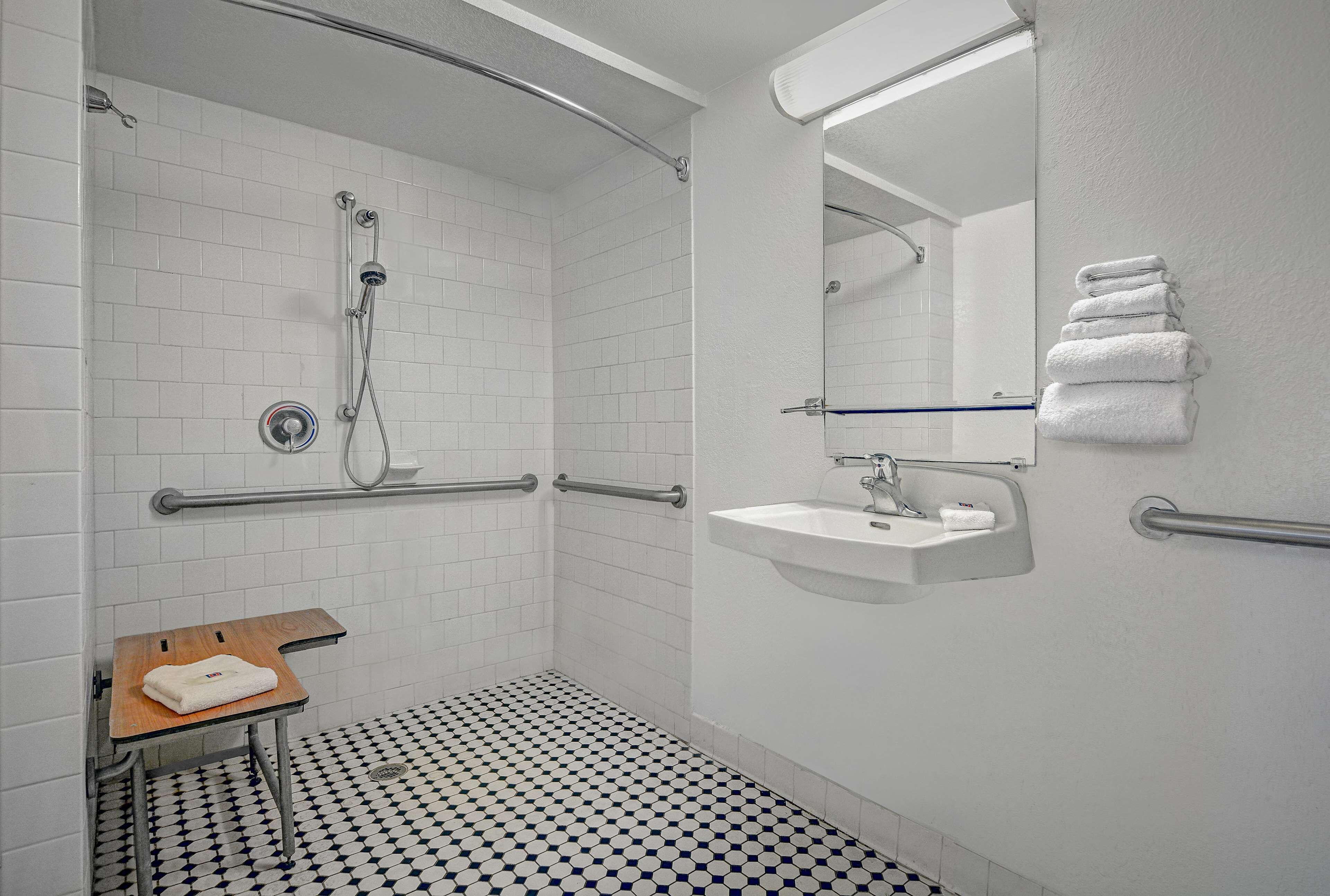 Baño con paredes de azulejos blancos, un lavamanos, un espejo y una ducha. Se observan barras de apoyo y un pequeño banco de madera.