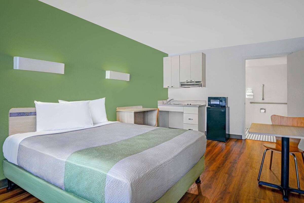 A bedroom with a bed, a desk, a chair, and a kitchenette.