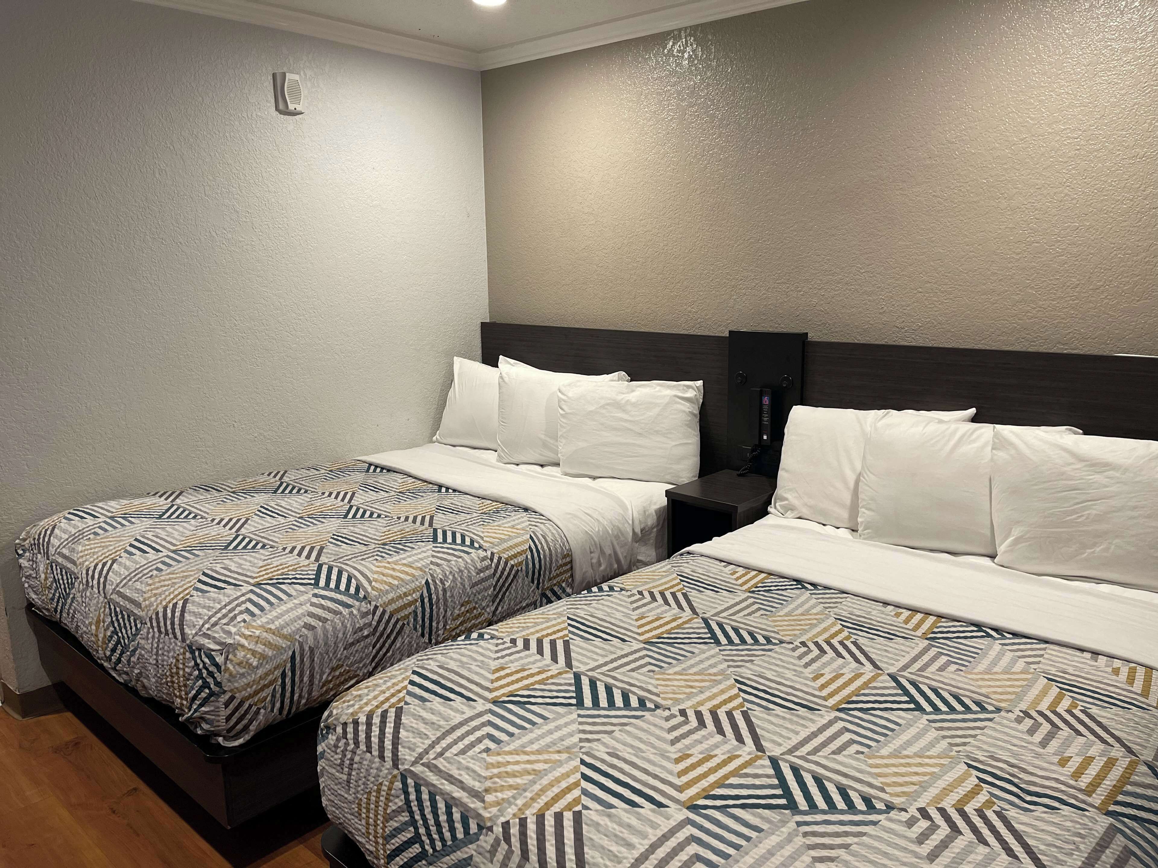 A hotel room with two beds, each with patterned bedding and white pillows. There is a bedside table with a phone and a small lamp. The walls are textured and the room has a wooden floor.