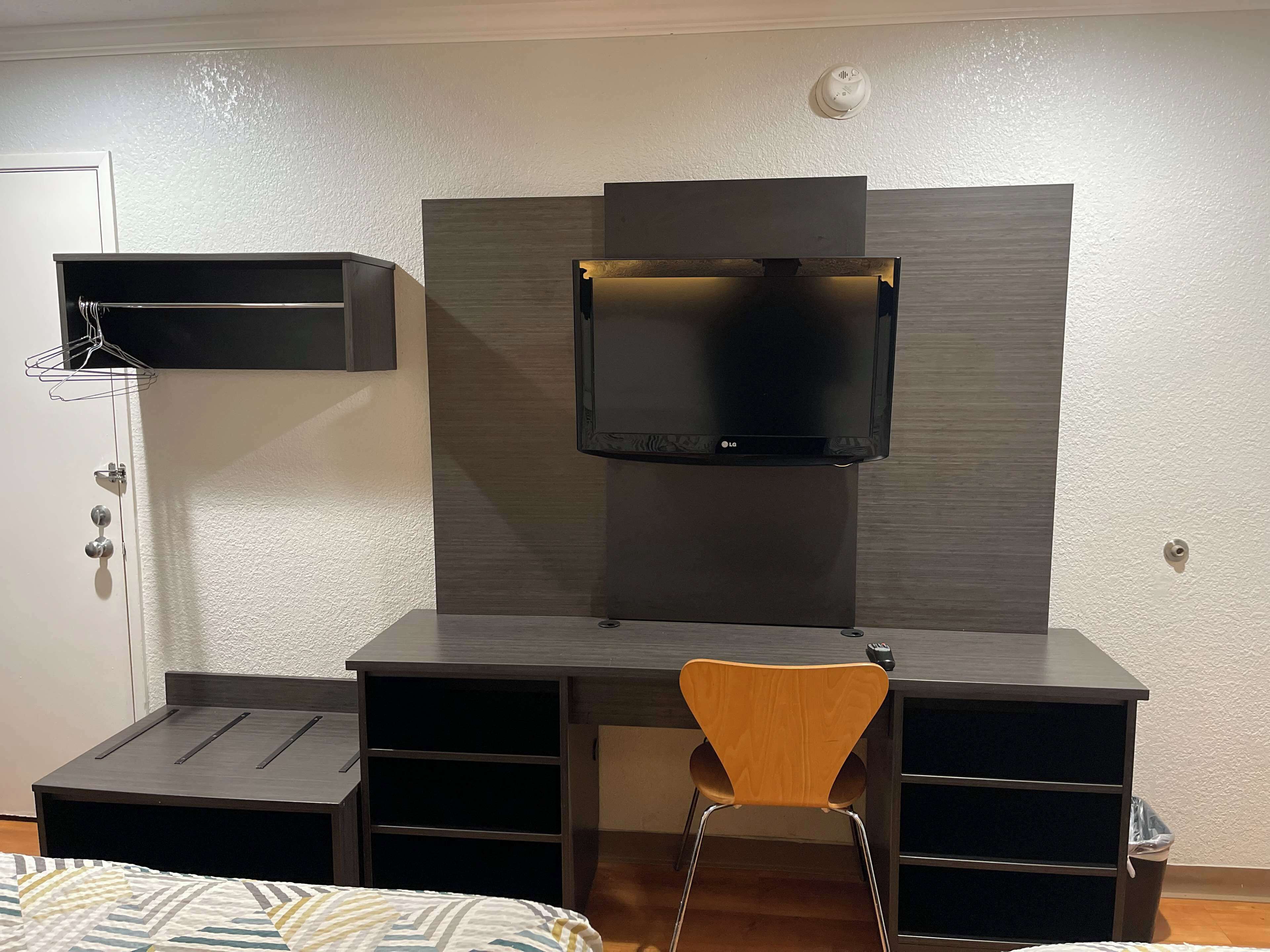 Une petite pièce comprenant un lit, un bureau avec une chaise, un téléviseur fixé au mur, un porte-manteau et une porte.