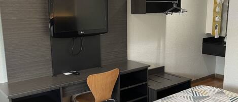 A modern hotel room with a bed, a desk with a chair, a TV mounted on the wall, and a small shelf with toiletries.