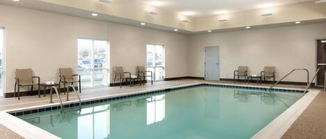 An indoor swimming pool with clear water, surrounded by beige chairs and a tiled floor.