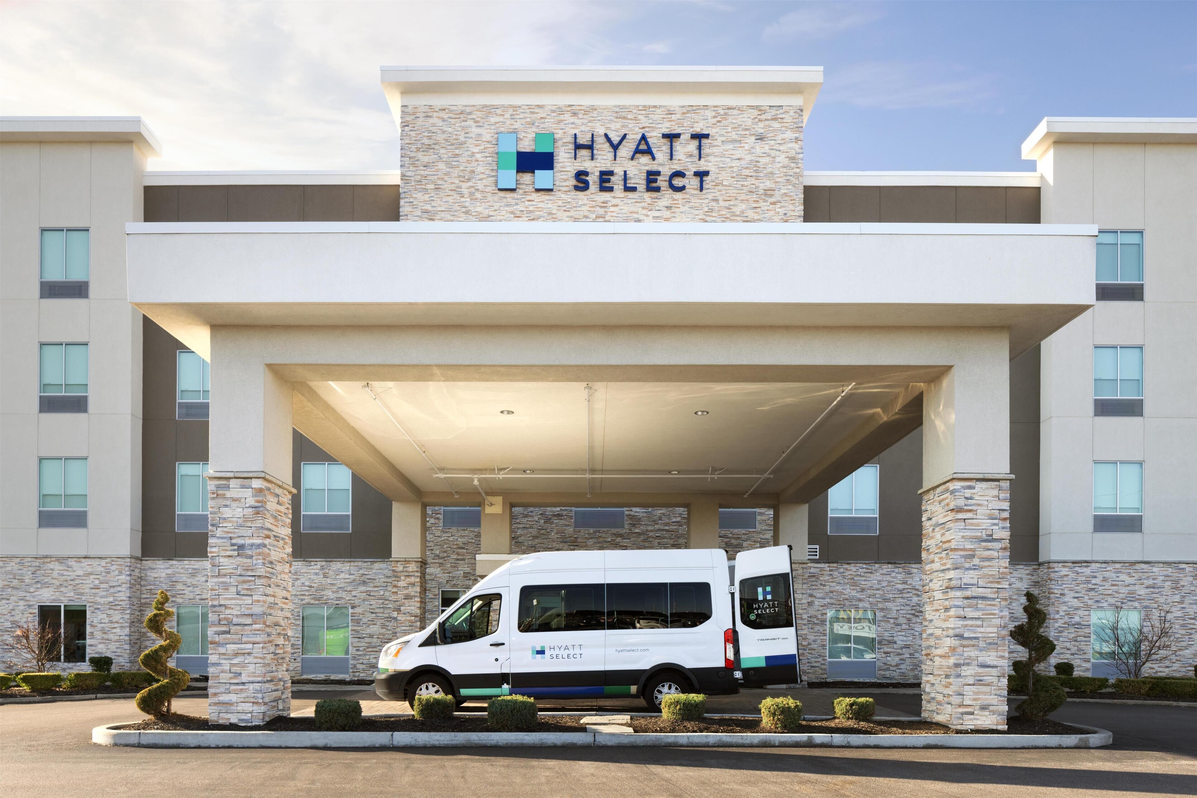 A modern hotel entrance with a white van parked in front, stone pillars, and a clear sky.