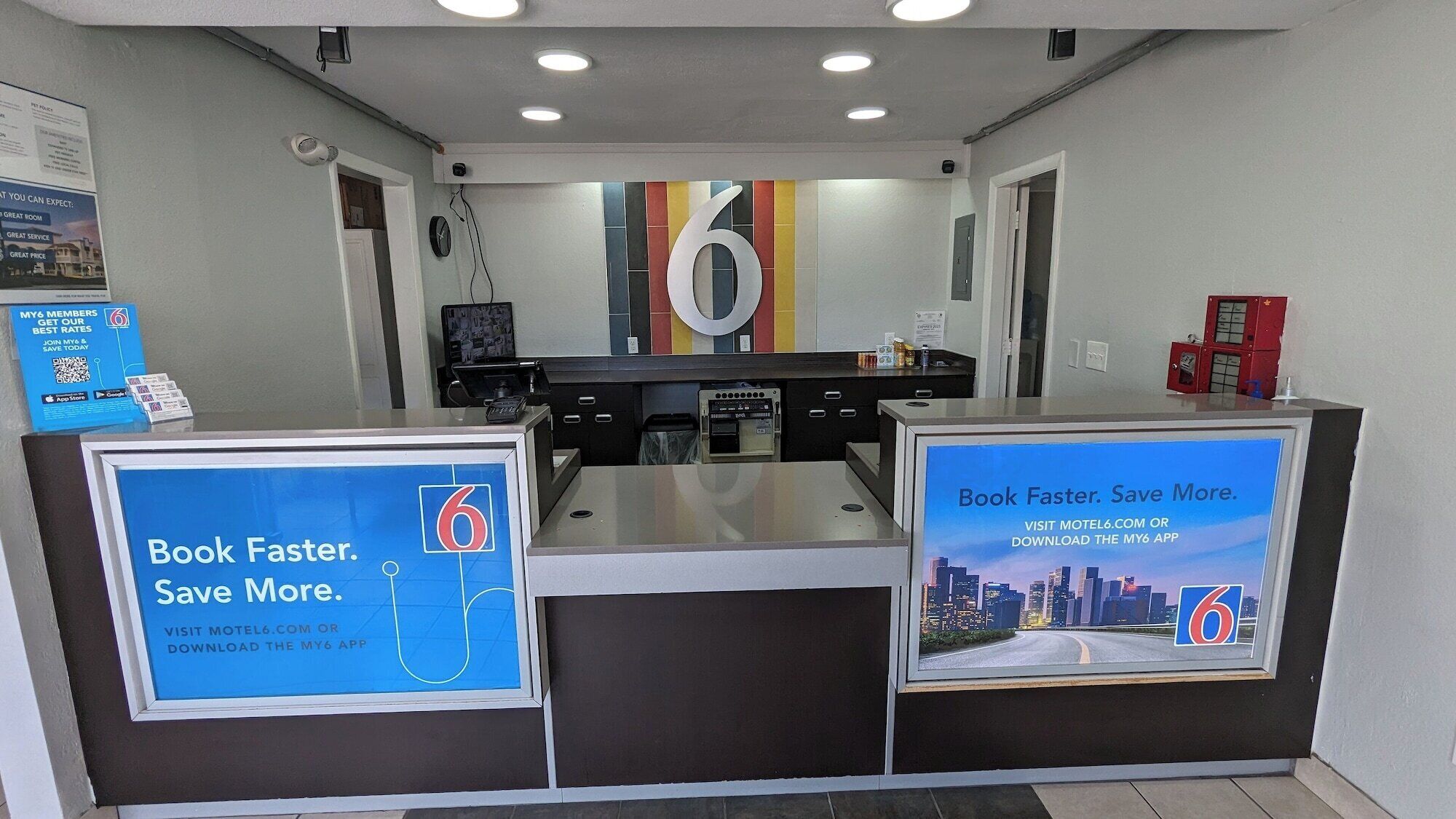 A reception desk with a large number '6' on the wall, two digital screens displaying promotional content, and a small trash bin.