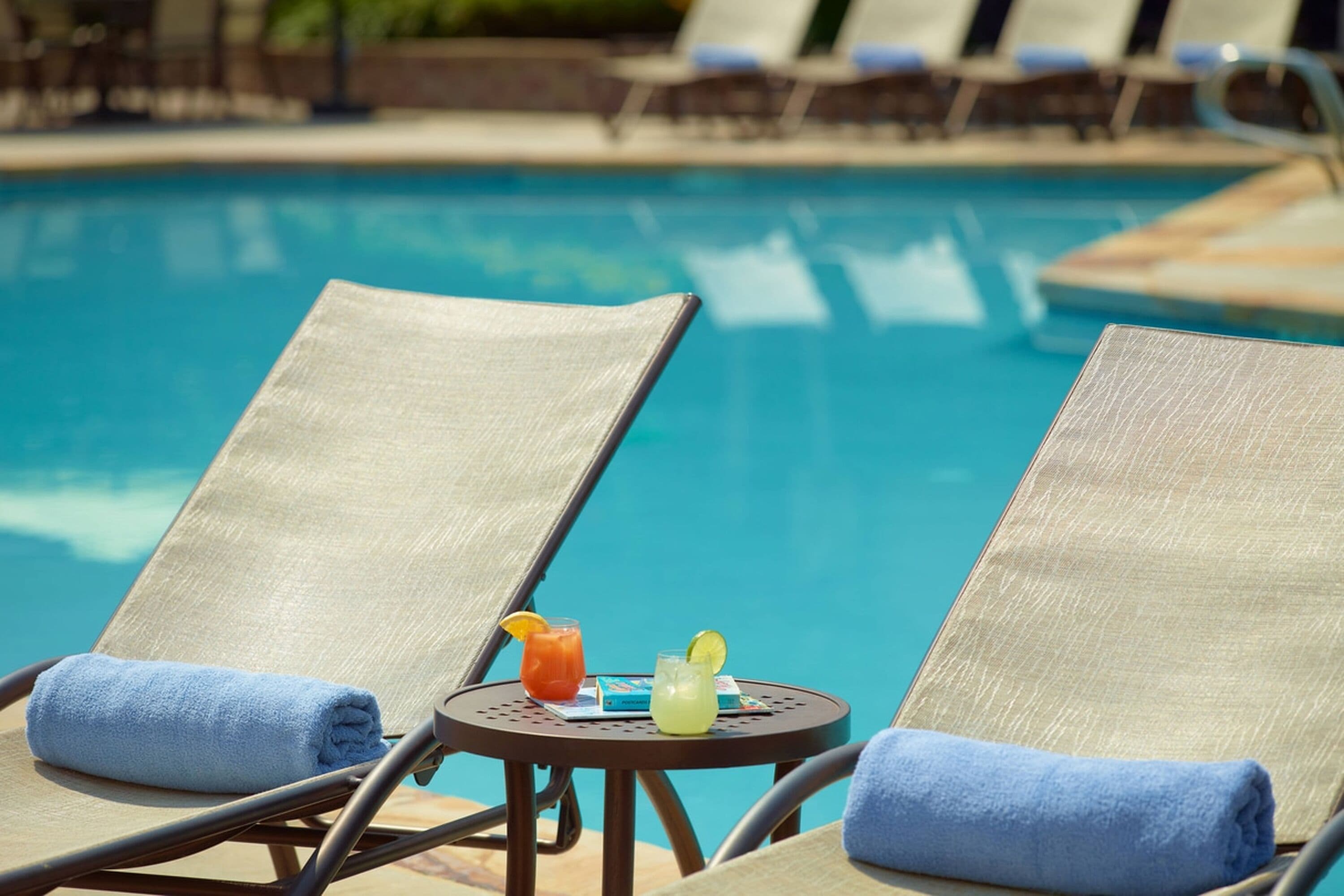 Two lounge chairs with blue towels, a small table with drinks, and a pool in the background.