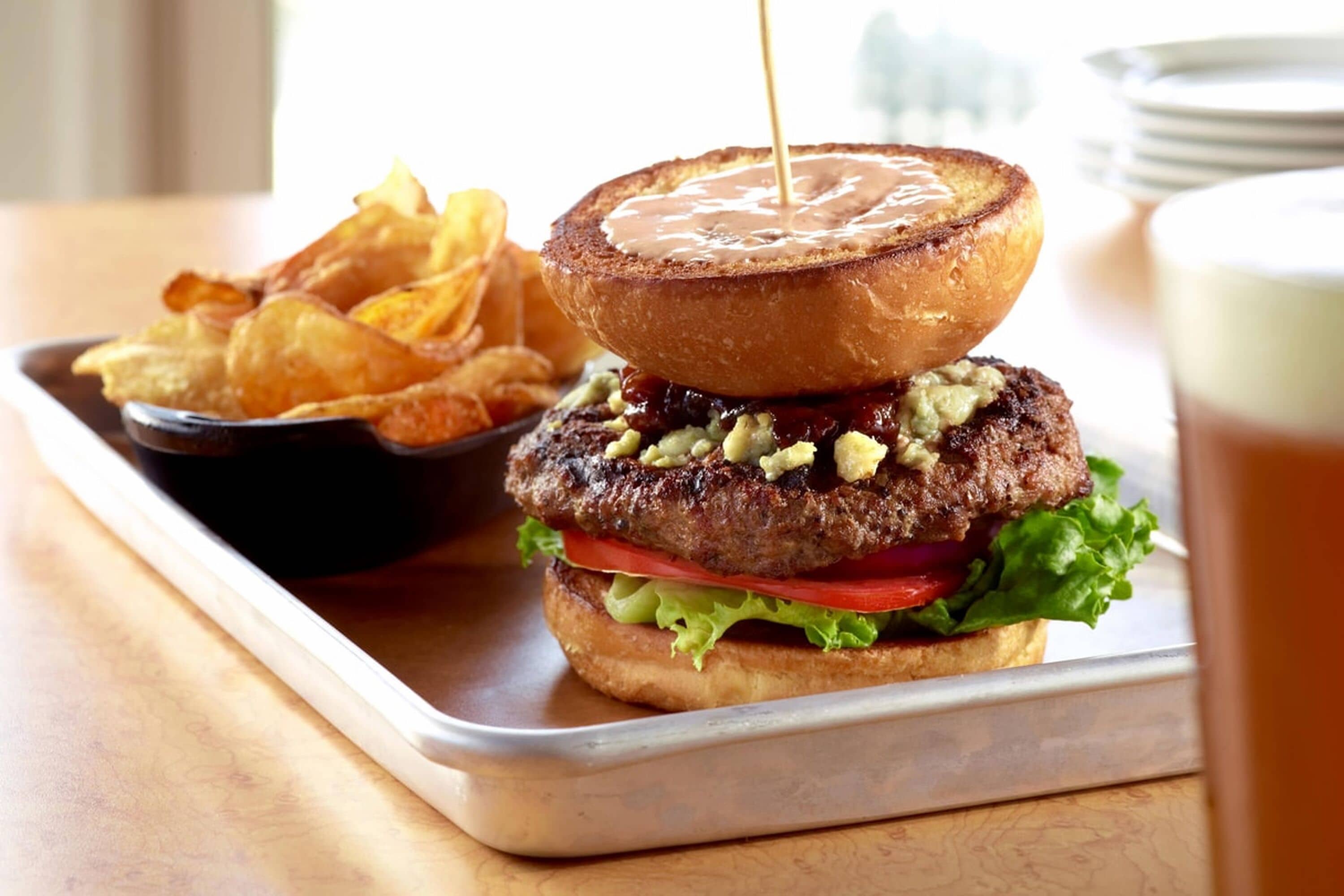 A burger with a toasted bun, lettuce, tomato, and a patty, served with chips and a drink.