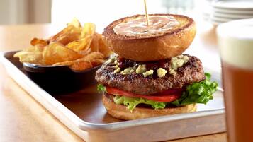 A burger with a toasted bun, lettuce, tomato, and a patty, served with chips and a drink.