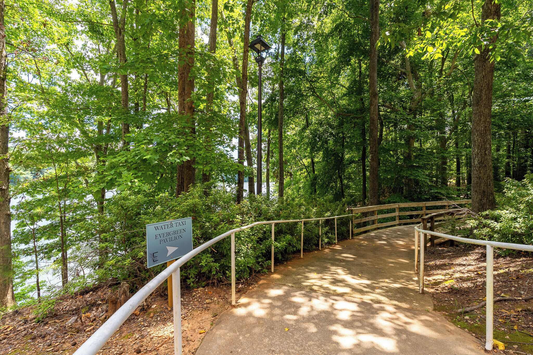 A concrete pathway with metal railings, surrounded by dense green foliage and tall trees. A sign indicates a water taxi and an everglade pavilion.