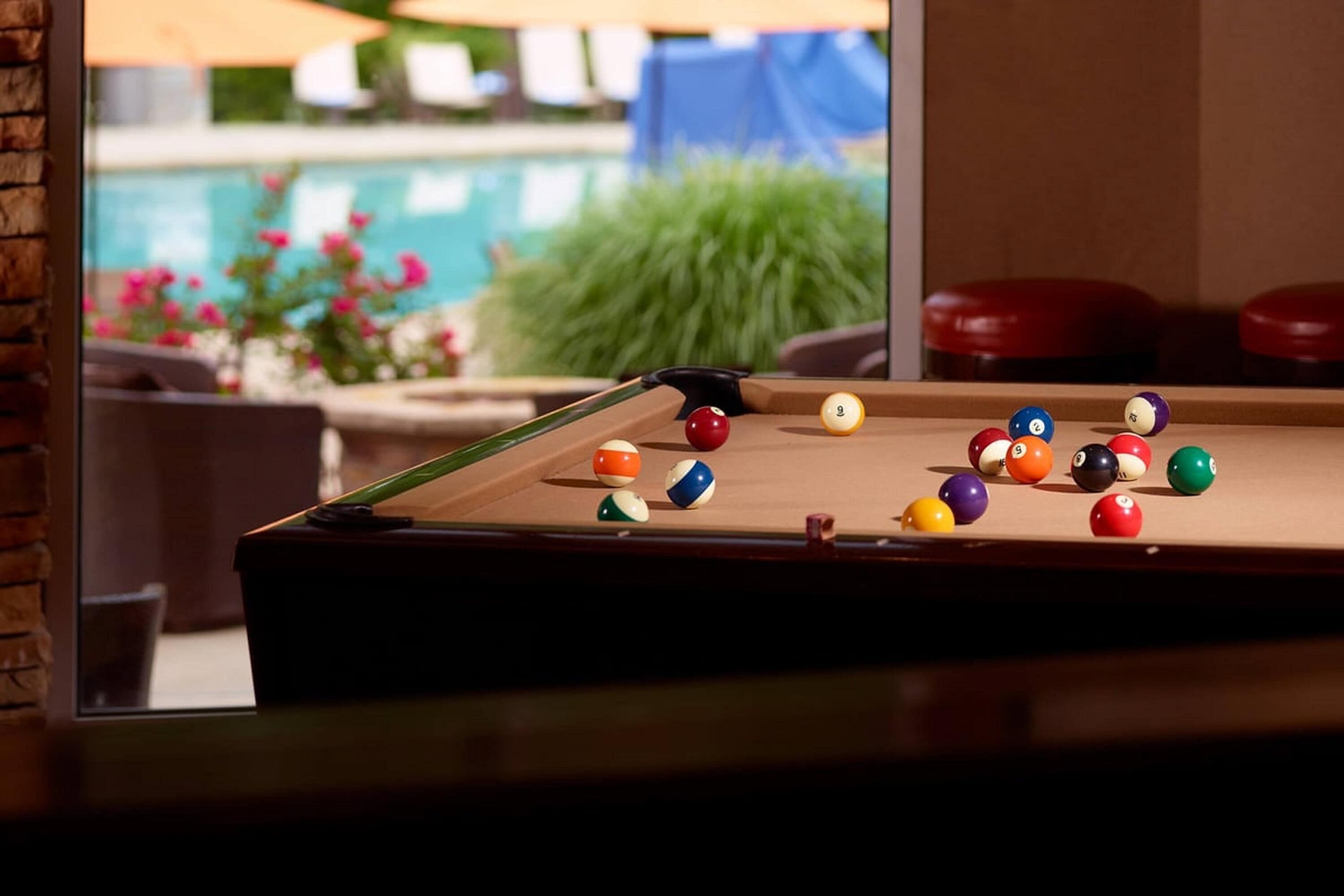 Uma mesa de bilhar com bolas, taco e vista pela janela para uma piscina e área de estar externa.