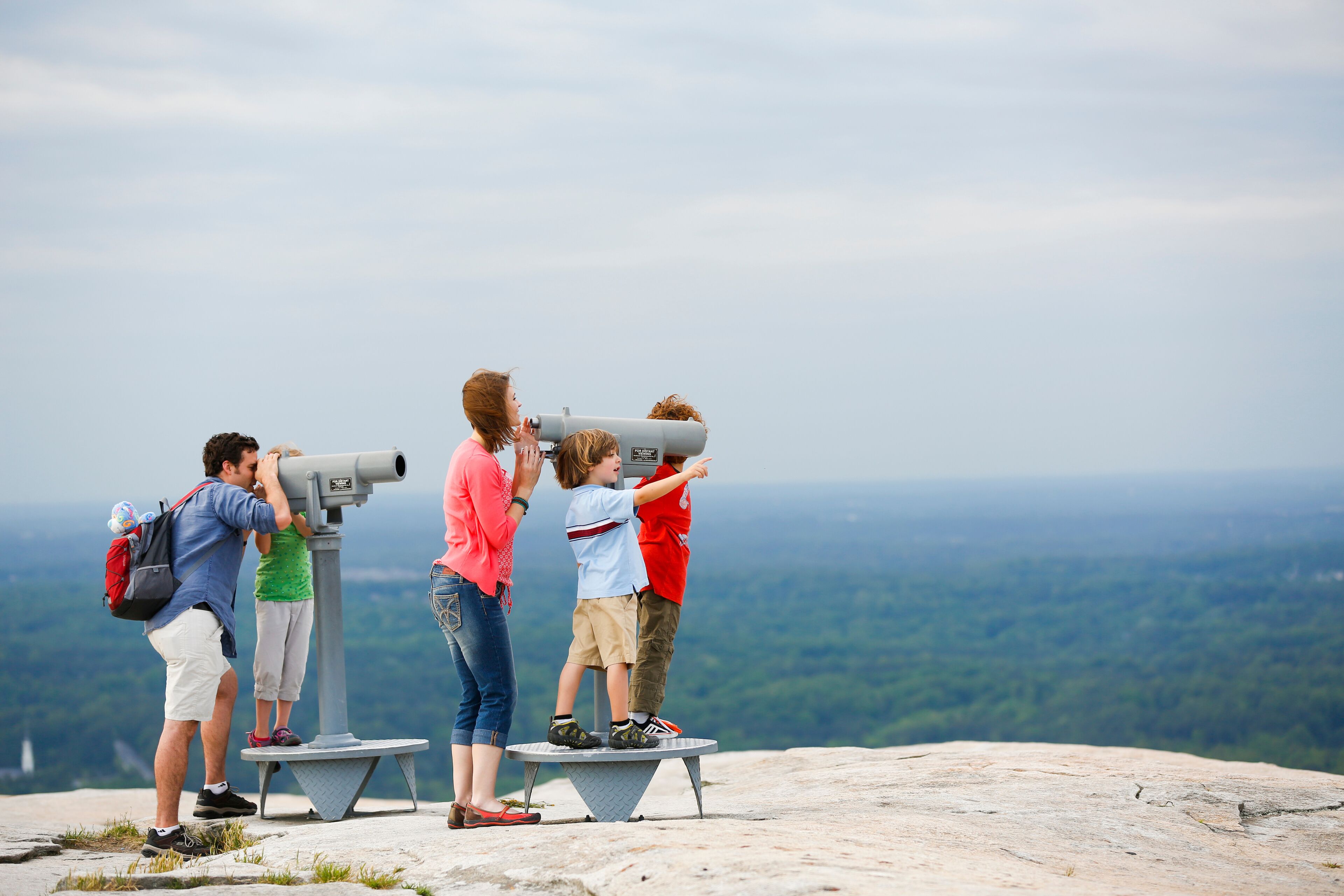 A group of people using telescopes on a rocky outcrop with a view of a forested landscape.