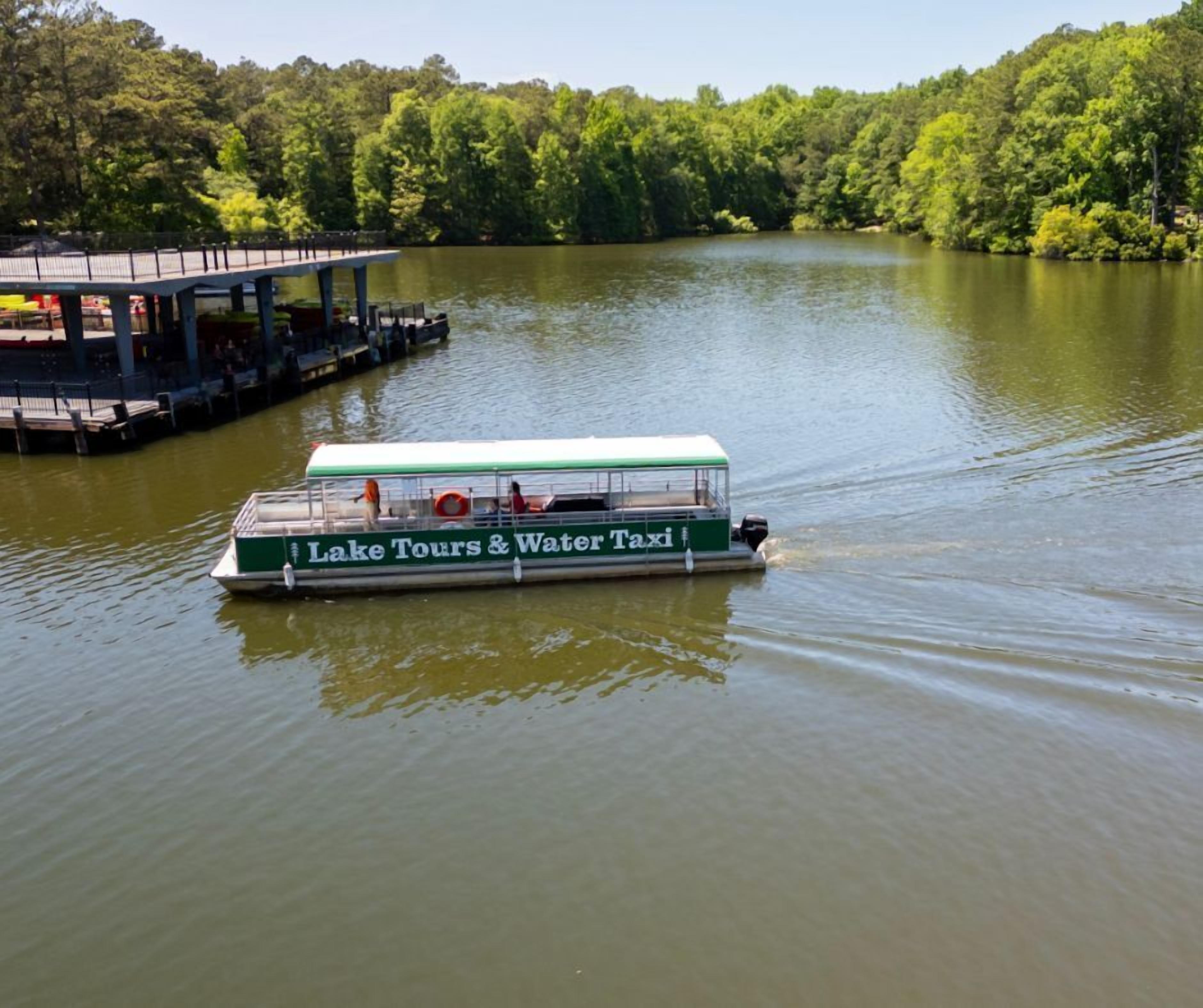 A boat with 'Lake Tours & Water Taxi' written on its side, floating on a lake with a dock and trees in the background.
