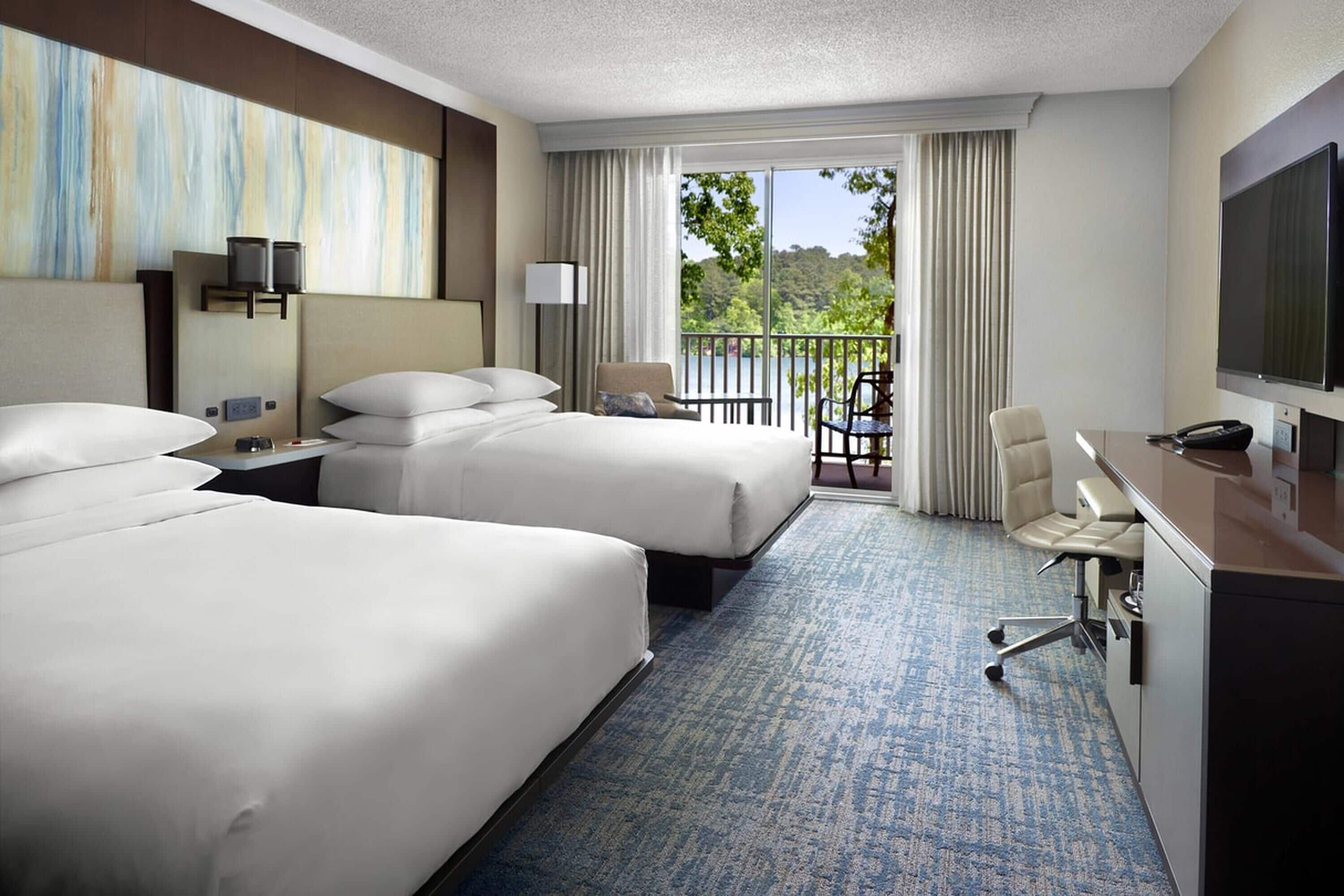 A hotel room with two beds, a desk, a chair, a TV, and a balcony with a view.