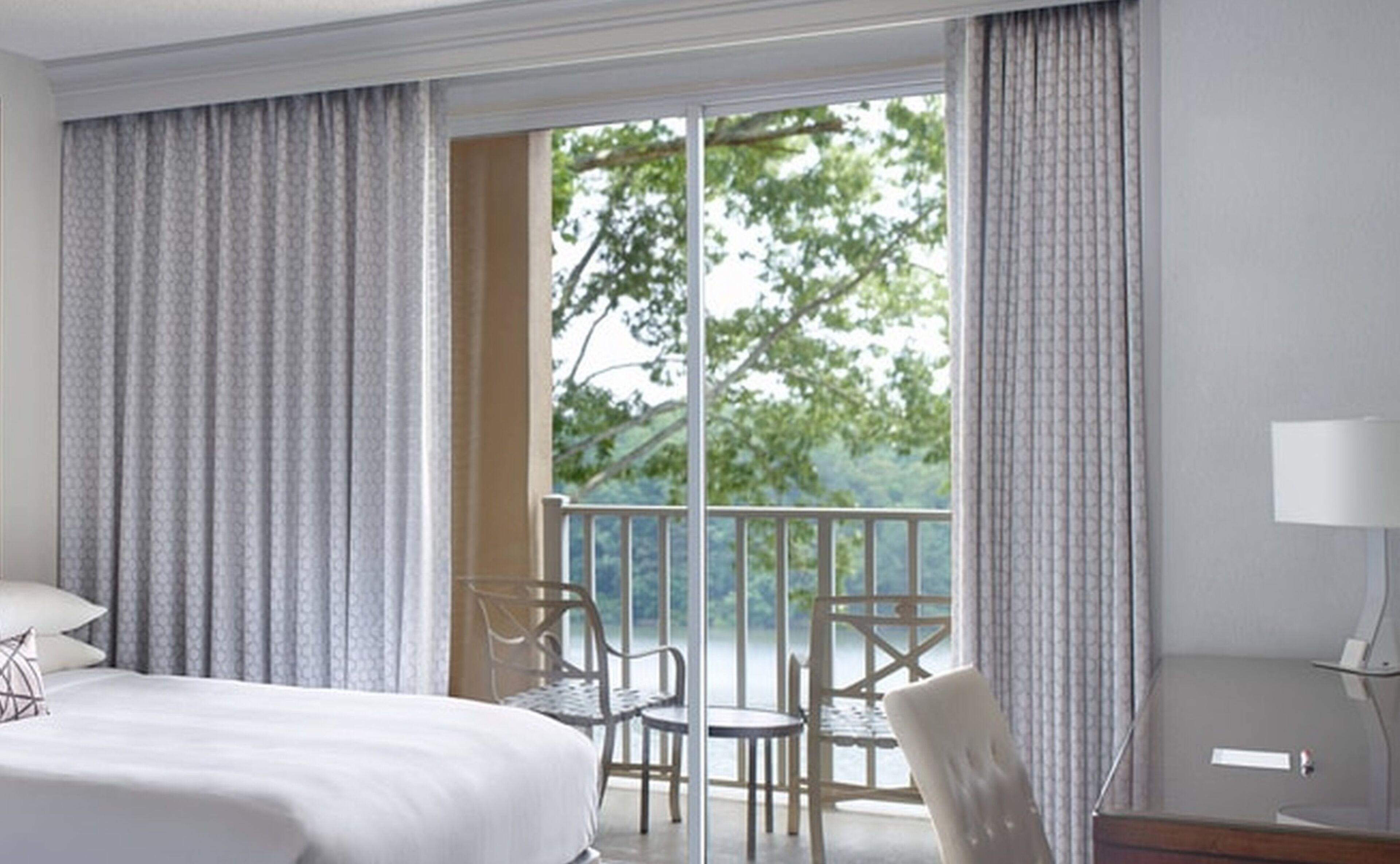 A hotel room with a large bed, a desk with a chair, and a balcony with a view of trees.