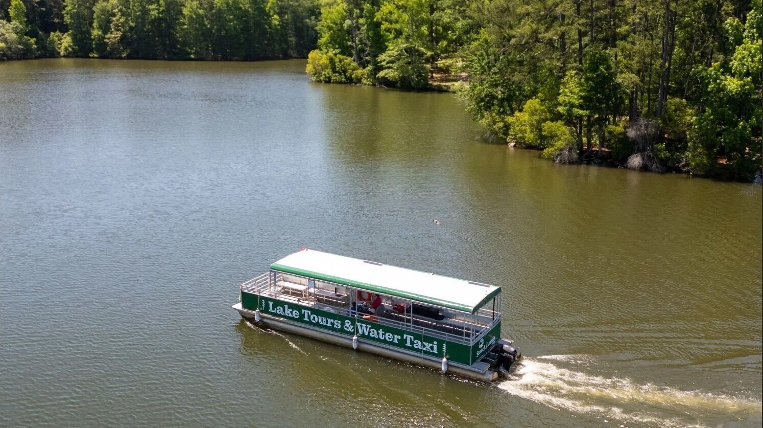 Un bateau vert et blanc, sur lequel on peut lire les mots « Excursions sur le lac et service de transport fluvial », se déplace sur un lac paisible, entouré d’une forêt dense.
